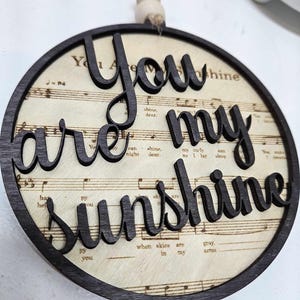 May include: A round wooden ornament with a black border and a white background featuring musical sheet music. The words "You are my sunshine" are cut out of the wood and are black.