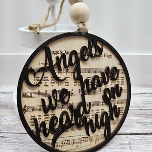 May include: A round wooden ornament with a brown background and a black outline. The ornament features a musical note background with the words "Angels We Have Heard On High" written in black cursive.