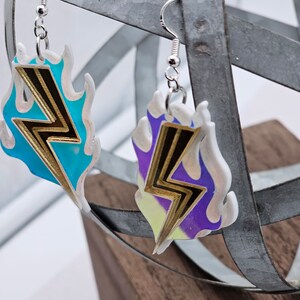 Könnte beinhalten: Ein Paar silberne Ohrringe mit einem Blitz-Design. Der Blitz ist golden und vor einem blauen und violetten Flammenhintergrund.
