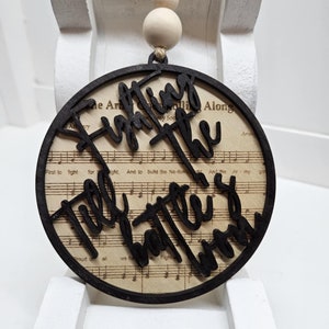 May include: A round wooden ornament with a black cutout design that reads "Fighting the Good Battle". The ornament is decorated with a sheet music background and has a twine hanger with wooden beads.
