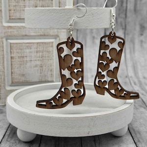 May include: A pair of brown wooden earrings shaped like cowboy boots with a heart pattern cut out. The earrings are hanging from silver hooks.