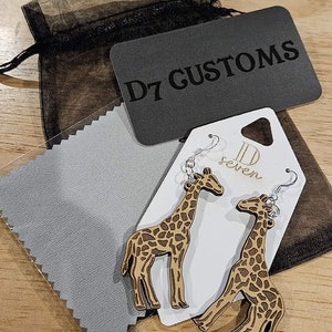 May include: A pair of wooden giraffe earrings with brown spots and silver hooks, presented on a white tag with "D seven" text. A black mesh bag, a grey cleaning cloth, and a black tag with "D7 CUSTOMS" are also visible.