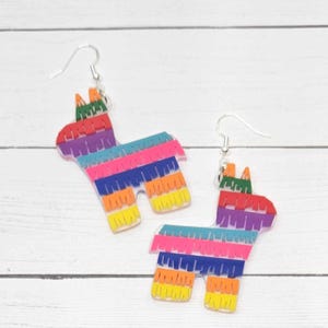 May include: A pair of colorful pinata earrings. Each earring is shaped like a donkey with a rainbow-colored fringe design. The earrings have silver-colored fish hook ear wires.