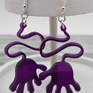 Peut inclure: Boucles d'oreilles en acrylique violet en forme de mains reli&eacute;es par une ligne ondul&eacute;e. Les boucles d'oreilles ont des crochets argent&eacute;s.