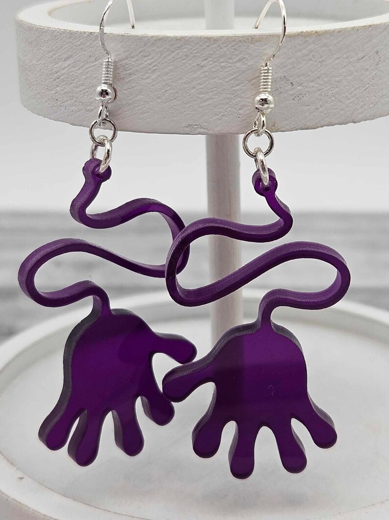 Peut inclure: Boucles d'oreilles en acrylique violet en forme de mains reli&eacute;es par une ligne ondul&eacute;e. Les boucles d'oreilles ont des crochets argent&eacute;s.