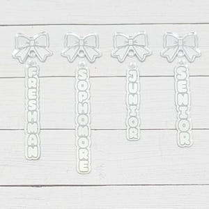 May include: Four clear acrylic charms, each featuring a bow at the top and a vertical tag below. The tags are engraved with the words FRESHMAN, SOPHOMORE, JUNIOR, and SENIOR in a bold, rounded font. Each charm includes a small hole for attachment.