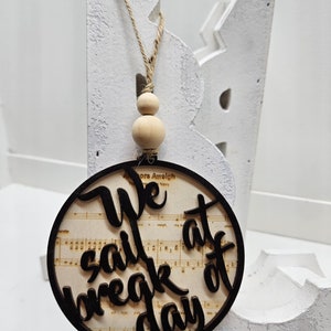 Puede incluir: Un adorno redondo de madera con un diseño recortado en negro que dice "We sail at break of day" sobre un fondo de partituras musicales antiguas. El adorno está colgado de una cuerda de yute con cuentas de madera.
