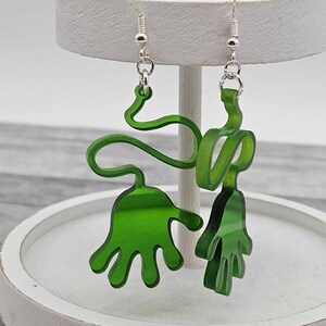 Peut inclure: Une paire de boucles d'oreilles en acrylique vertes en forme de mains. Les boucles d'oreilles sont suspendues &agrave; des crochets argent&eacute;s.