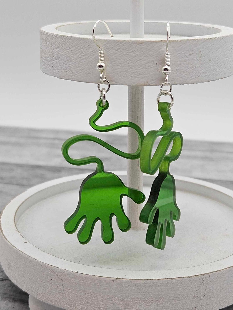 Peut inclure: Une paire de boucles d'oreilles en acrylique vertes en forme de mains. Les boucles d'oreilles sont suspendues &agrave; des crochets argent&eacute;s.