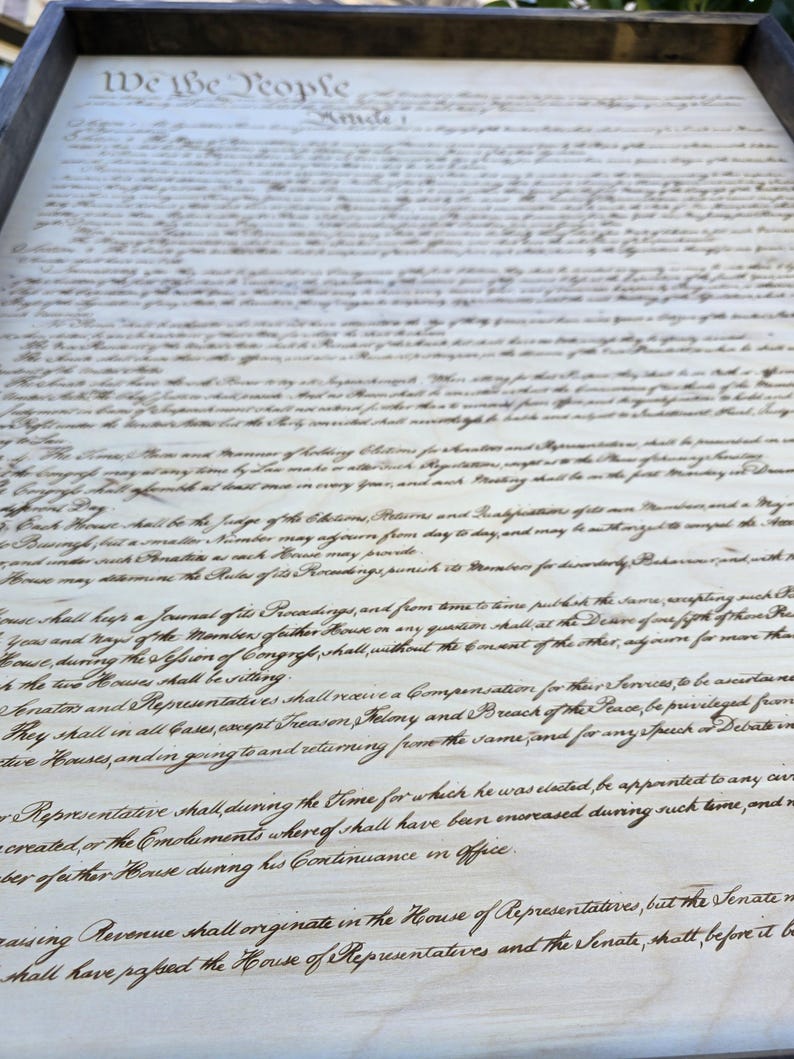 May include: A close-up of a wood-burned image of the US Constitution. The text "We the People" is visible at the top of the document. The document is framed in a dark wood frame.