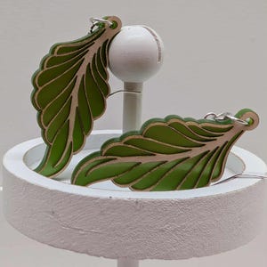 May include: A pair of leaf-shaped earrings in green and tan, with a detailed vein pattern. The wooden earrings are displayed on a white stand, featuring a white ball and a circular white base.