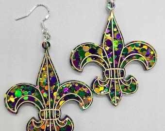 May include: A pair of gold-colored fleur-de-lis earrings with green, purple, and gold glitter accents. The earrings have silver hooks.