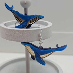 May include: A pair of blue whale-shaped earrings with gold accents. The earrings are displayed on a white stand. The whales have detailed fins and a decorative pattern on the side. The earrings are suspended from silver-colored hooks.