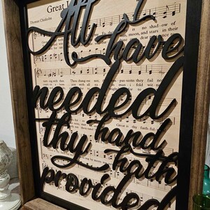 May include: Black wooden sign with the words "All I have needed thy hand hath provided" in a decorative font. The sign is framed and has a background of sheet music.
