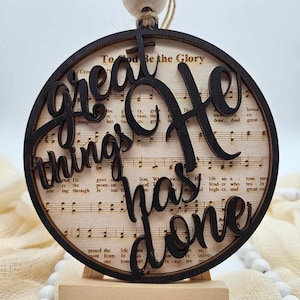 May include: A round wooden ornament with a black background and white music notes. The words "Great things He has done" are cut out of the wood in a black script font.