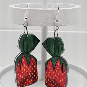 May include: A pair of dangle earrings featuring a red and green strawberry design. The earrings are made of acrylic and have silver hooks.