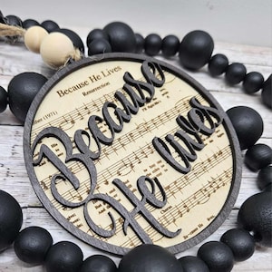 以下が含まれることがあります： 楽譜の背景に「Because He Lives」という言葉を筆記体で書いた丸い木製の看板。黒いビーズのガーランドから吊り下げられている。