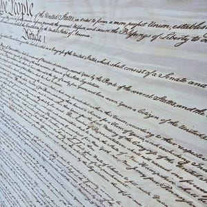 May include: A close-up of a wood panel with the first paragraph of the United States Constitution, written in cursive script. The text reads, "We the People of the United States, in Order to form a more perfect Union, establish Justice, insure domestic Tranquility, provide for the common defence, promote the general Welfare, and secure the Blessings of Liberty to ourselves and our Posterity, do ordain and establish this Constitution for the United States of America."