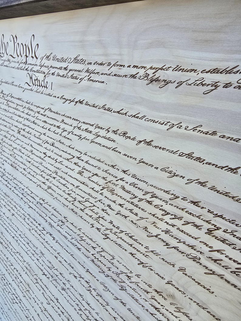 May include: A close-up of a wood panel with the first paragraph of the United States Constitution, written in cursive script. The text reads, "We the People of the United States, in Order to form a more perfect Union, establish Justice, insure domestic Tranquility, provide for the common defence, promote the general Welfare, and secure the Blessings of Liberty to ourselves and our Posterity, do ordain and establish this Constitution for the United States of America."