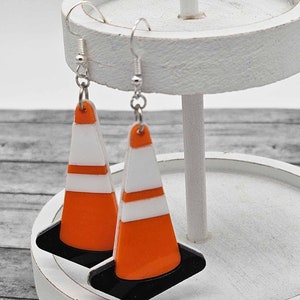 May include: A pair of dangle earrings featuring orange and white traffic cone designs. The cones have a black base and are attached to silver hooks.