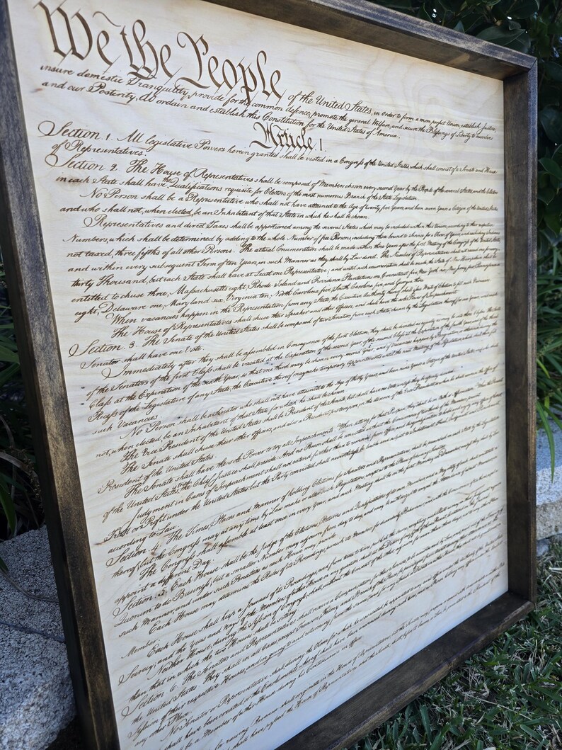May include: A framed wood print of the United States Constitution. The text is in black ink on a white background. The frame is dark brown wood.