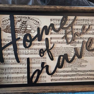 May include: A wooden framed sign with a black background and a white sheet of music with the words "Home of the Brave" cut out in black and layered on top. The sheet music is from the song "The Star-Spangled Banner".