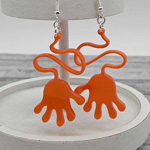 Peut inclure: Boucles d'oreilles en acrylique orange en forme de mains avec un design ondul&eacute;. Les boucles d'oreilles sont suspendues &agrave; des crochets argent&eacute;s.