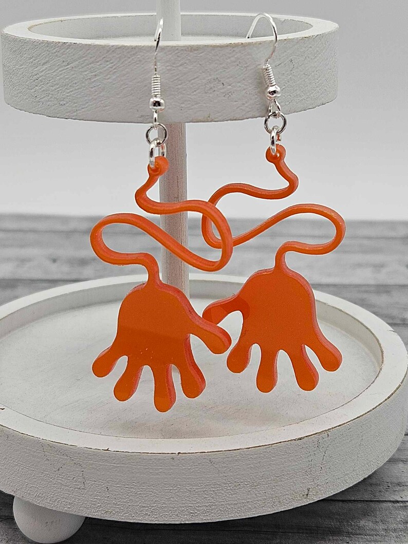 Peut inclure: Boucles d'oreilles en acrylique orange en forme de mains avec un design ondul&eacute;. Les boucles d'oreilles sont suspendues &agrave; des crochets argent&eacute;s.