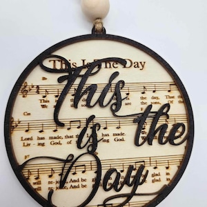 May include: A round wooden ornament with a black background and the words "This is the Day" in a decorative font. The background is a sheet of music.
