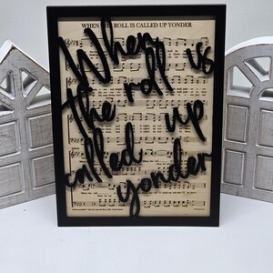 Puede incluir: Un letrero negro enmarcado con fondo blanco que presenta una pieza de arte de palabras en cursiva negra en 3D que dice "When the roll is called up yonder". El fondo es una hoja de música.