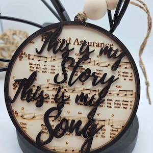 May include: A round wooden ornament with a black border and a white background. The ornament features a musical score with the words "This is my Story This is my Song" written in black cursive.