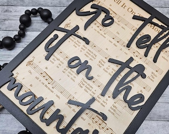 May include: Black wooden sign with the words "Go tell it on the mountain" cut out in the shape of the words. The sign is framed with a black frame and has a vintage sheet music background.