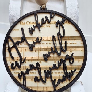 May include: A round wooden ornament with a black border and the words "Where we will always know" written in cursive on a background of sheet music.