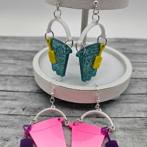 May include: A pair of novelty earrings featuring a bucket design. One pair is teal with yellow shovels, the other is pink with purple shovels. Both pairs have white handles and silver earring hooks.