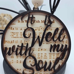 以下が含まれることがあります： 「It Is Well With My Soul」という文字が黒くくり抜かれた丸い木製オーナメント。楽譜の背景と、上部に白いビーズが飾られています。