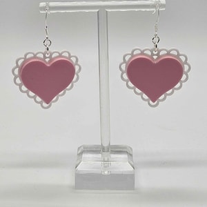 May include: A pair of pink heart-shaped earrings with a white lace trim. The earrings are hanging from silver hooks.
