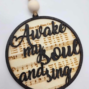 May include: A round wooden ornament with the words "Awake my soul and sing" cut out in black script. The background features sheet music. The ornament is attached to a string with wooden beads.