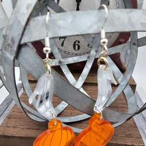May include: A pair of dangle earrings featuring a clear plastic pumpkin with an orange pumpkin charm. The earrings are hanging from a silver hook.