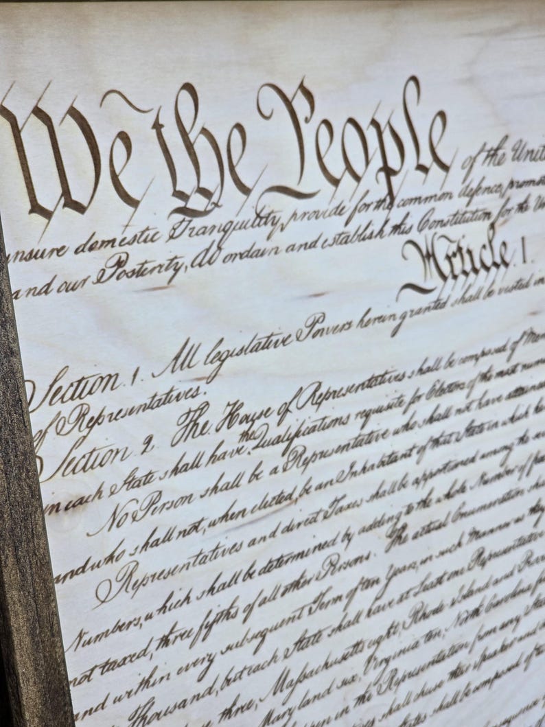 May include: Close-up view of a wood-framed print featuring the preamble to the United States Constitution. The text, 'We the People', is prominently displayed in elegant calligraphy. The document's aged appearance is suggested by the color and texture of the background.  The entire piece is presented in a rustic, antique style, suitable for historical or patriotic settings.  The detailed script of the Constitution's text is clearly visible.