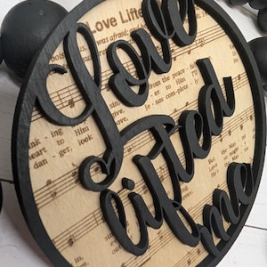 May include: A circular wooden decoration with the phrase "Love Lifted Me" in black script. The background shows sheet music, and the ornament has a black border. A jute string is attached for hanging.