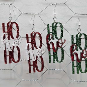 May include: Three pairs of dangle earrings with the word "Ho Ho Ho" written on them. The earrings are made of glitter and come in red, green, and white.