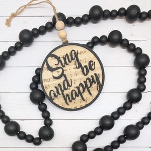 May include: A wooden bead garland featuring a round wooden ornament. The ornament is inscribed with the words "Sing and be happy" and set against a musical score. The beads are black, and the ornament is suspended by twine.