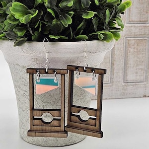 Puede incluir: Un par de pendientes de madera con forma de guillotina con un panel translúcido rosa y azul. Los pendientes cuelgan de ganchos plateados.