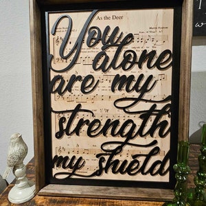Może przedstawiać: Oprawiona grafika z nutami w tle i słowami "You alone are my strength my shield" w czarnym skrypcie. Rama jest ciemnobrązowa z czarną obwódką. Tło jest jasno-beżowe, tworząc efekt warstwowy.