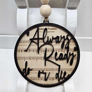 May include: A round wooden ornament with a black border and a vintage sheet music background. The ornament features the text "Always Ready to or Die" in black script.
