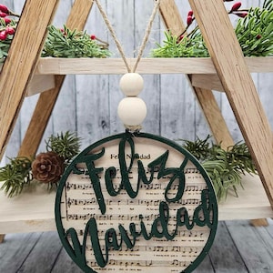 May include: A green wooden ornament with the words "Feliz Navidad" cut out of the wood. The ornament is decorated with a sheet music background.