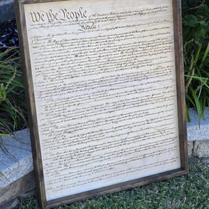 May include: A framed wood print of the United States Constitution. The document is written in cursive script and includes the phrase "We the People".