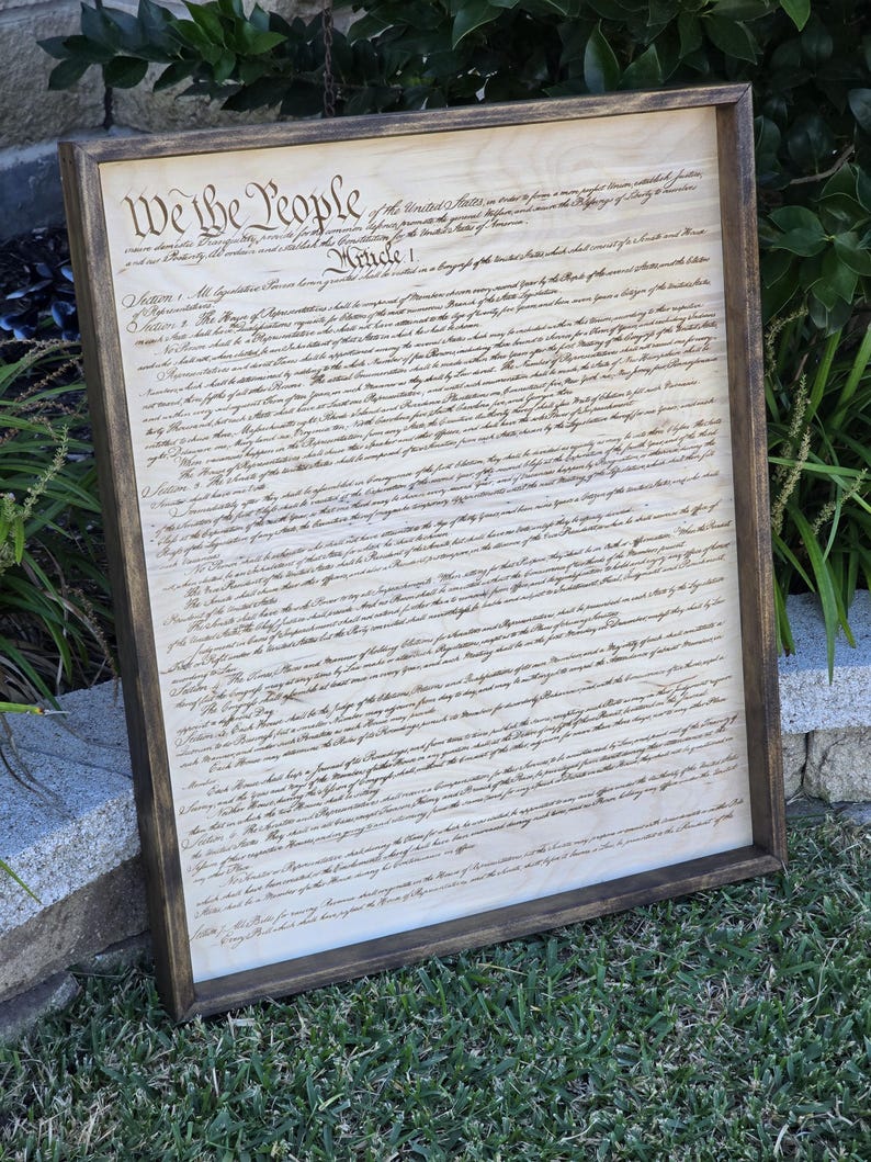 May include: A framed wood print of the United States Constitution. The document is written in cursive script and includes the phrase "We the People".