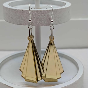 May include: Gold-coloured, geometric earrings with a fan-like design. The earrings hang from silver-coloured hooks and are displayed on a white, circular stand. The earrings have a layered, Art Deco style, reminiscent of the 1920s.