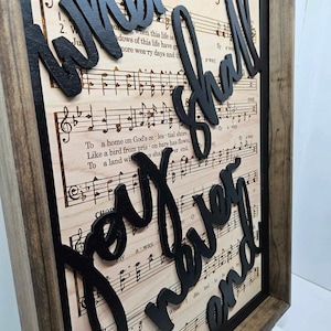 May include: A wooden framed wall art piece with a black background and white sheet music. The words "When I Shall  Wander On" are written in black, 3D letters on top of the sheet music.